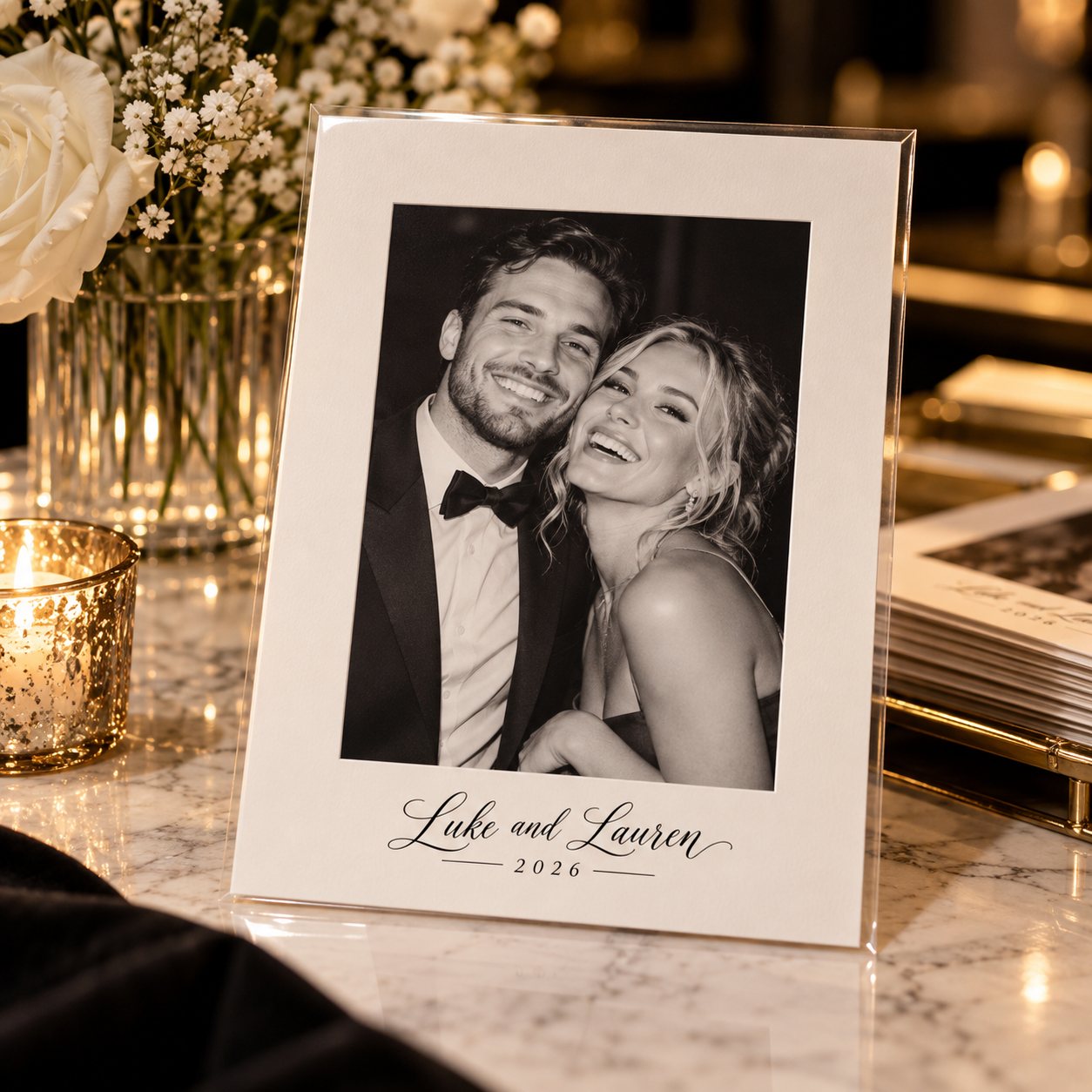 A black-and-white portrait of a couple on white cardstock printed with “Luke and Lauren 2026”, in a clear sleeve on a marble table